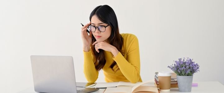 A doubtful and focused Asian female student in eyeglasses is looking at her laptop screen